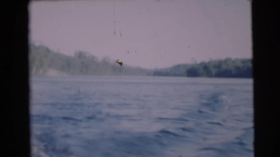 1953 8mm view from a moving boat near Baraboo, Wisconsin, showing rippling water, pale sky, and wooded shoreline.
