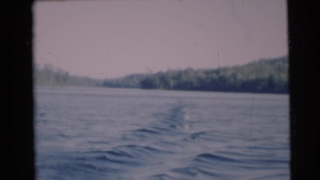 1953 home-movie frame from a moving boat near Baraboo, Wisconsin, with blue water ripples and a tree-lined shore in soft focus.