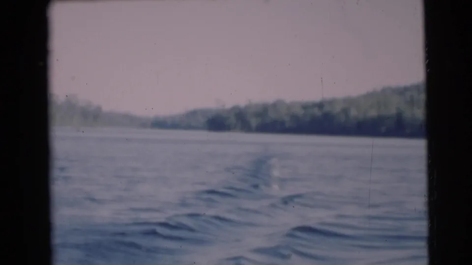1953 home-movie frame from a moving boat near Baraboo, Wisconsin, with blue water ripples and a tree-lined shore in soft focus.
