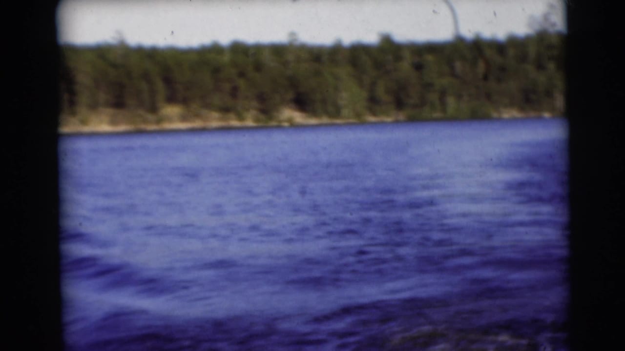1953 Baraboo, Wisconsin lake view with blue water, a wooded shore, pale sky, and visible 8mm film edges.