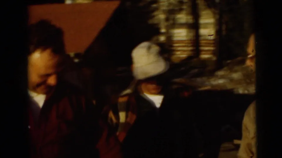 Later home-movie frame showing two partially visible adults in outdoor clothing near a rustic lakeside or camp area.