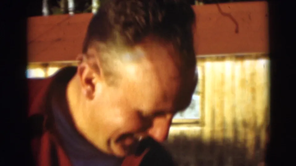 In 1953 Baraboo home-movie footage, an adult man in plaid outerwear smiles near a sunlit rustic exterior.