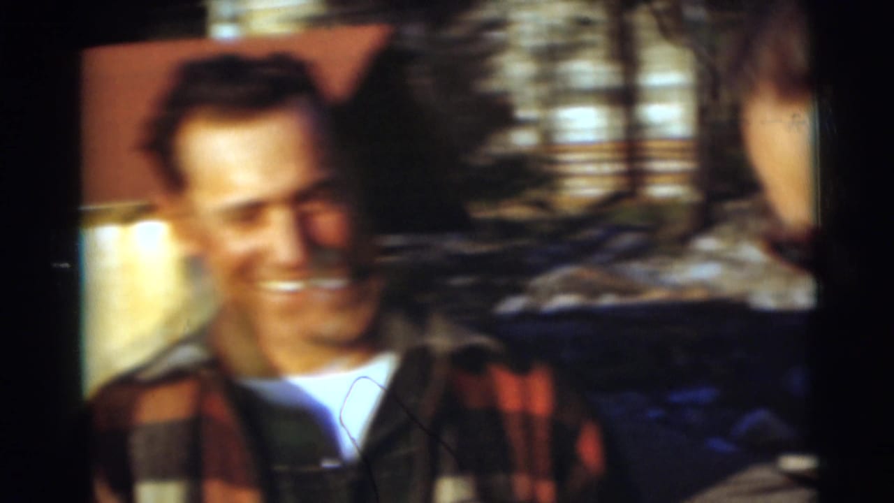 Smiling man in plaid outerwear stands partly obscured beside another figure outdoors.
