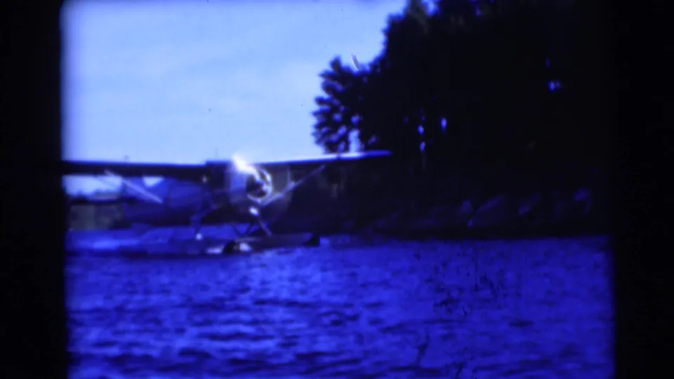 1953 Baraboo, Wisconsin home-movie frame showing a small floatplane on a lake beside trees, with dark film borders and blue color cast.