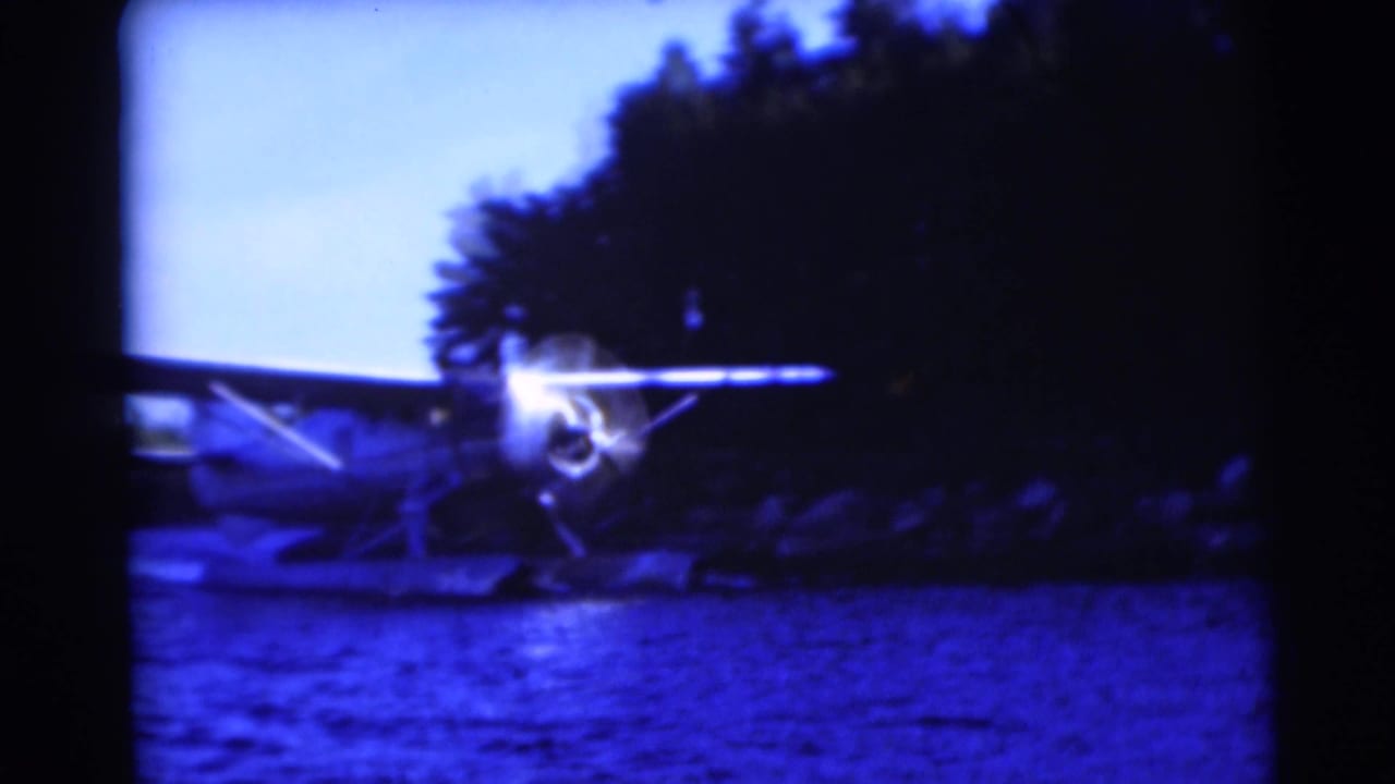 Blue-tinted 8mm frame of a small floatplane on a Baraboo lake, with a bright propeller area and trees behind it.