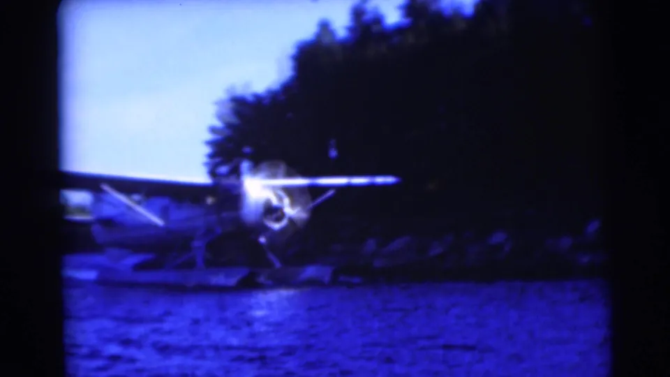 Blue-tinted 8mm frame of a small floatplane on a Baraboo lake, with a bright propeller area and trees behind it.