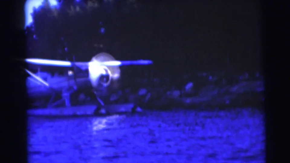 1953 home-movie frame showing the floatplane nearer the camera on lake water, with soft focus, blue cast, and a wooded bank behind.