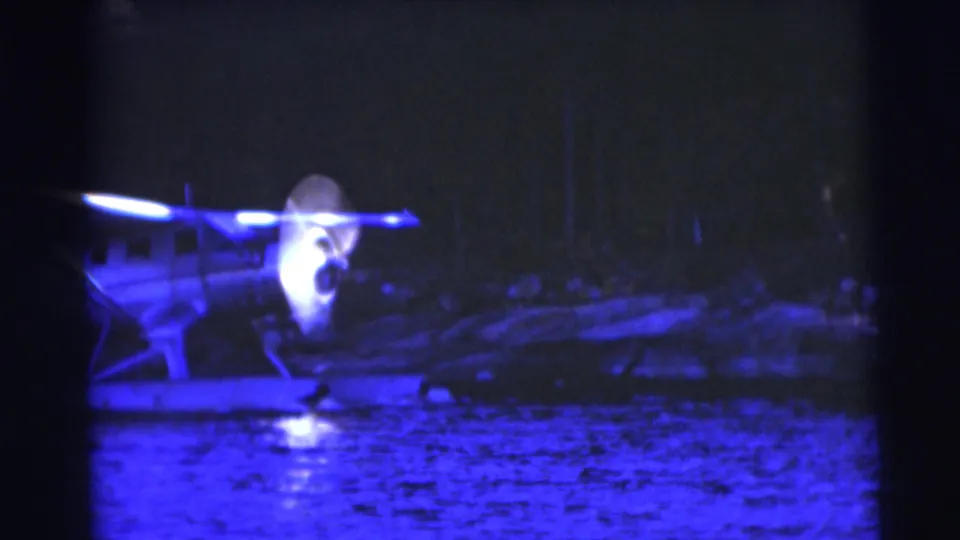 Blue-tinted 8mm frame of the floatplane on the lake near Baraboo, Wisconsin, with water reflections and a shaded wooded shore.