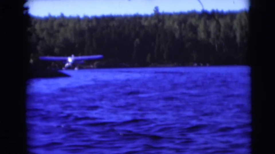 1953 Baraboo, Wisconsin 8mm frame showing a floatplane on rippled blue water with a dark wooded shoreline beyond.
