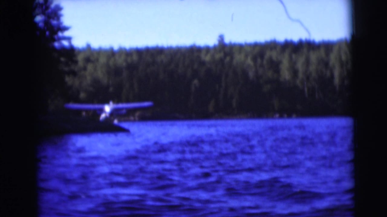1953 Baraboo, Wisconsin home-movie frame showing a small floatplane low over rippled lake water near a forested shore.