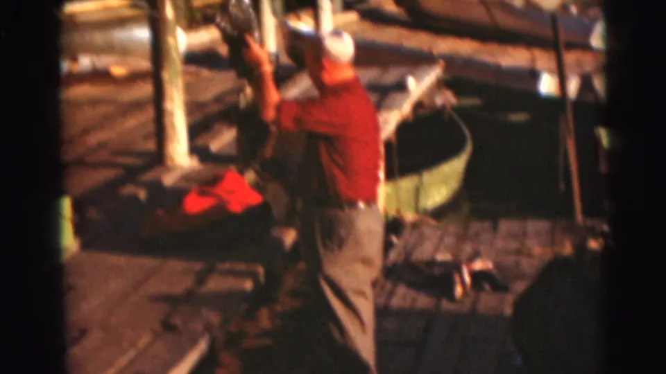 An adult wearing a red shirt and cap works on a wooden dock near boats in the 1953 Baraboo, Wisconsin lake footage.