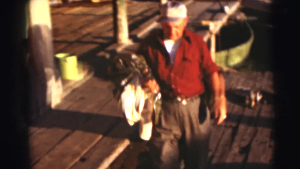 Adult man in a red shirt and cap walks along a Baraboo dock in 1953 carrying fishing gear and a white cloth.