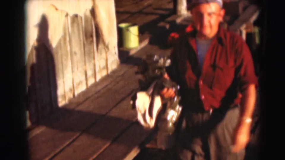 1953 dockside frame of an adult man in a red shirt approaching the camera with fishing gear and a white cloth in hand.