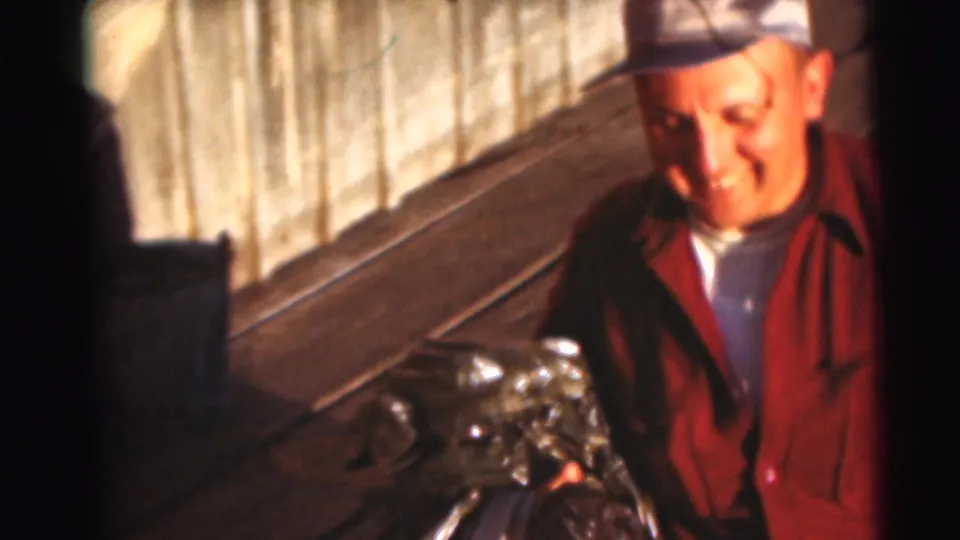 Close 1953 Baraboo home-movie view of a smiling adult man in a red shirt and cap beside fishing gear on a wooden dock.