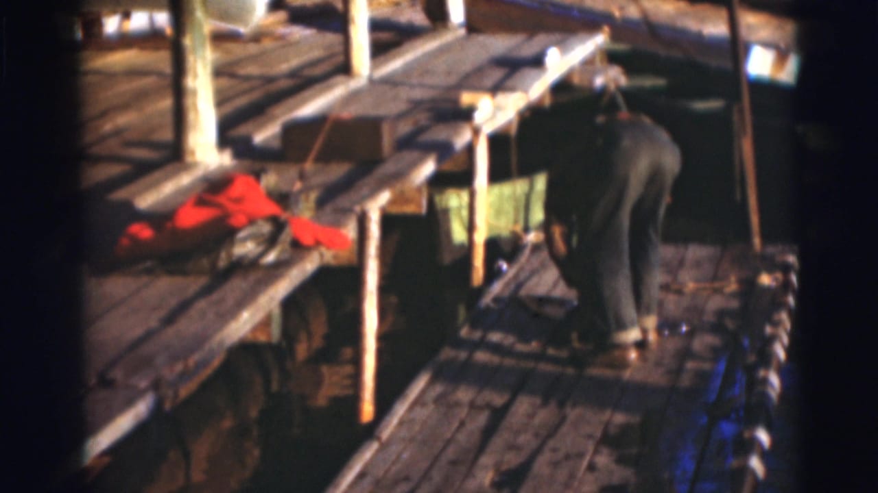 In 1953 Baraboo, Wisconsin footage, a man bends on a sunlit wooden dock near a red jacket and water.