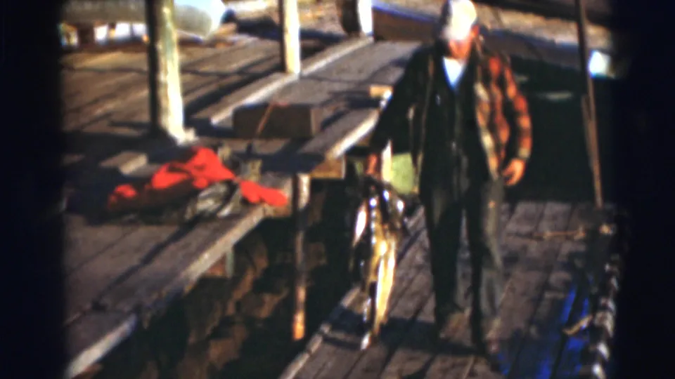 A man in a white hat stands on the sunlit dock, appearing to hold a fish as he turns toward the camera.