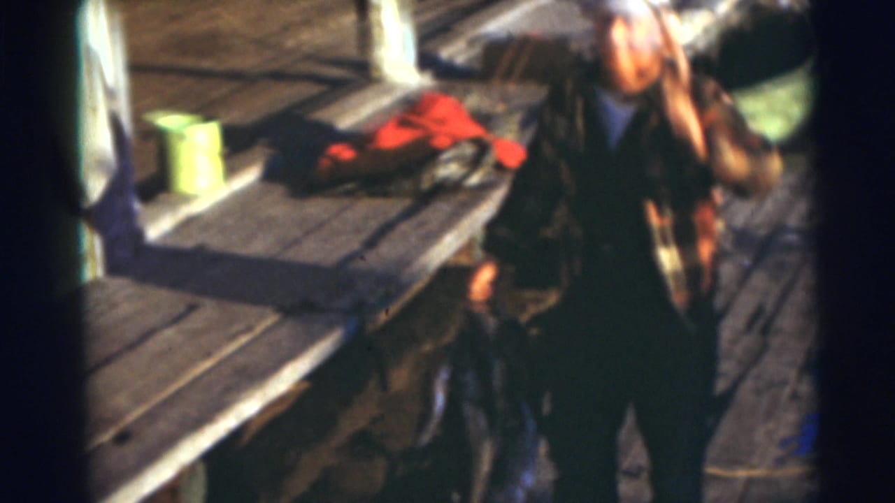 In 1953 Baraboo, Wisconsin footage, a man in a cap and plaid shirt stands near a wooden dock with fishing items nearby.