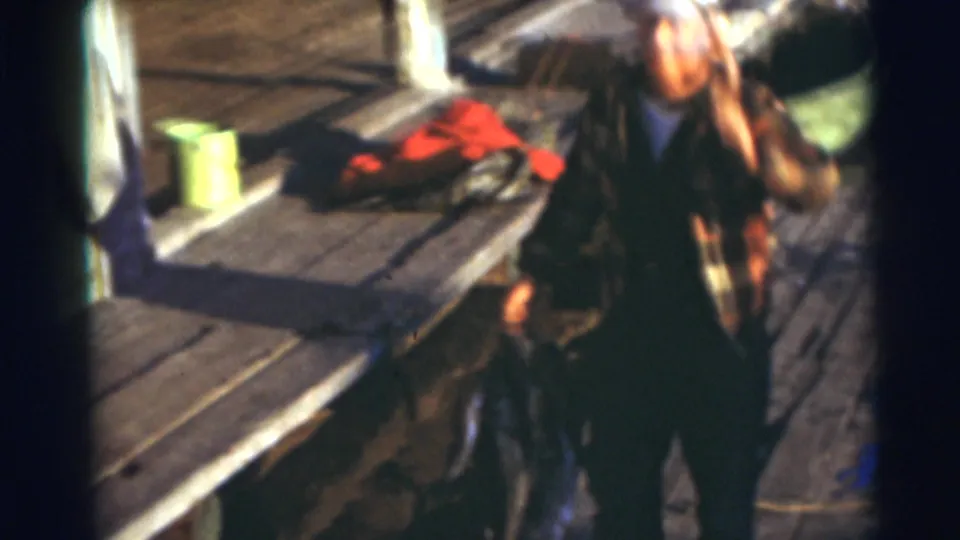 In 1953 Baraboo, Wisconsin footage, a man in a cap and plaid shirt stands near a wooden dock with fishing items nearby.