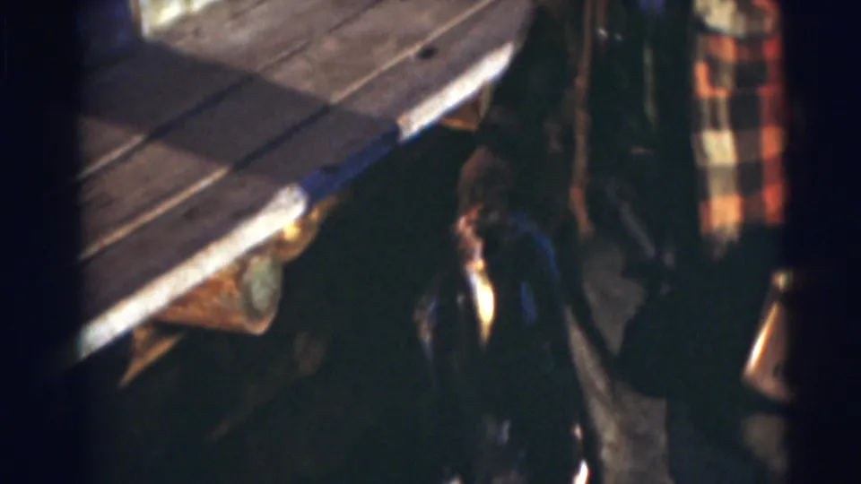 The home-movie frame shows the man near the wooden dock, with a fish and gear visible in the lower part of the shot.
