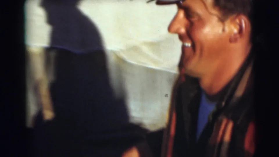 Close-up 1953 home-movie frame of the man smiling in profile, wearing a cap and plaid outer shirt after the fishing moment.