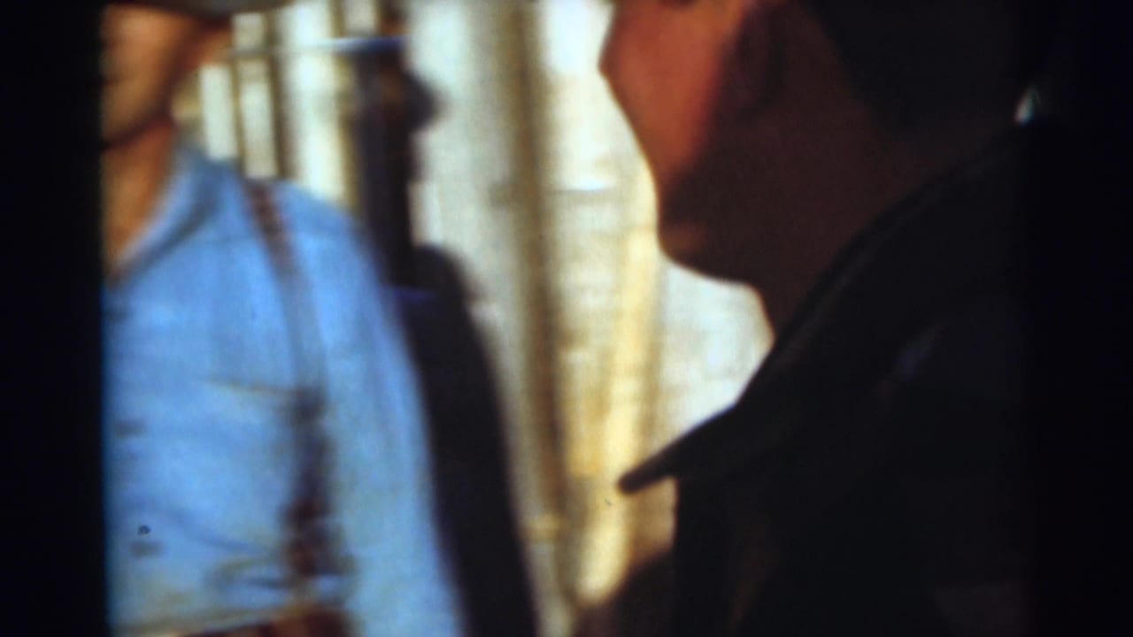1953 Baraboo home-movie frame showing an adult in a plaid shirt near camera with another shadowed figure in a dim setting.