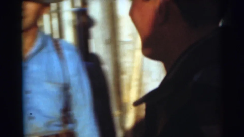 1953 Baraboo home-movie frame showing an adult in a plaid shirt near camera with another shadowed figure in a dim setting.