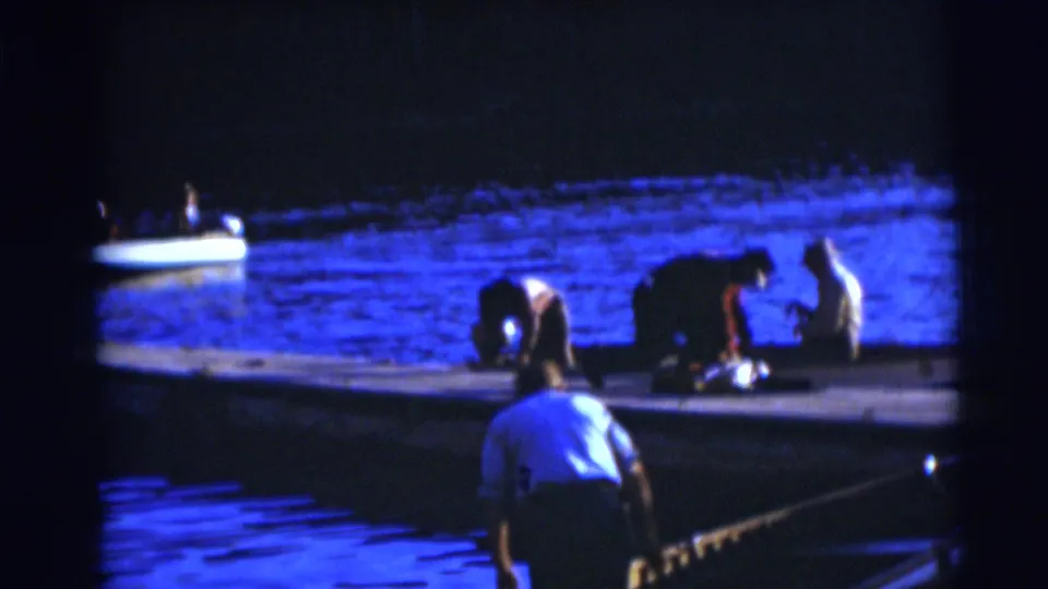 Nighttime Baraboo dock scene from 1953 with several people bending near the shoreline and a small boat on the water.