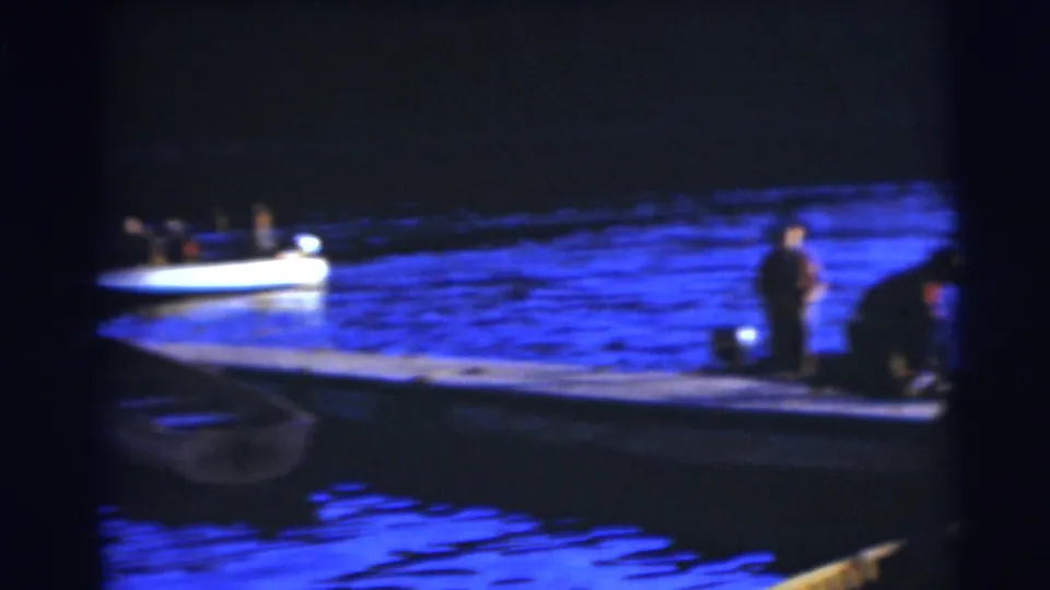 1953 Baraboo lakeside frame showing a small boat on blue-tinted water and people working near the dock at night.