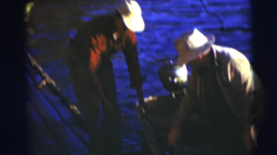 1953 Baraboo, Wisconsin home-movie frame showing two men in brimmed hats preparing around a rowboat beside the lake.