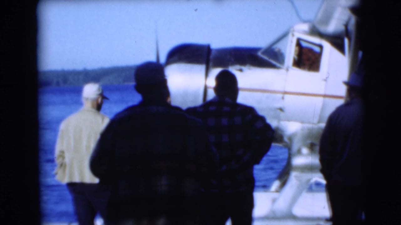 In Baraboo, Wisconsin, men in 1950s attire watch beside a white floatplane on calm water.