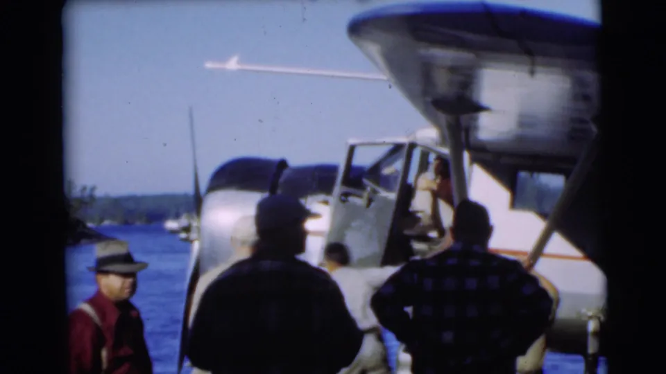 The camera frames men on the dock beneath the wing of a small white seaplane on a sunny Wisconsin lake.
