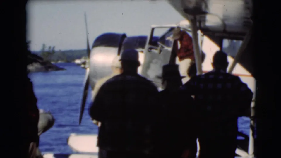 Men watch a person at the open door of a white floatplane during a 1953 lakeside boarding scene in Baraboo.