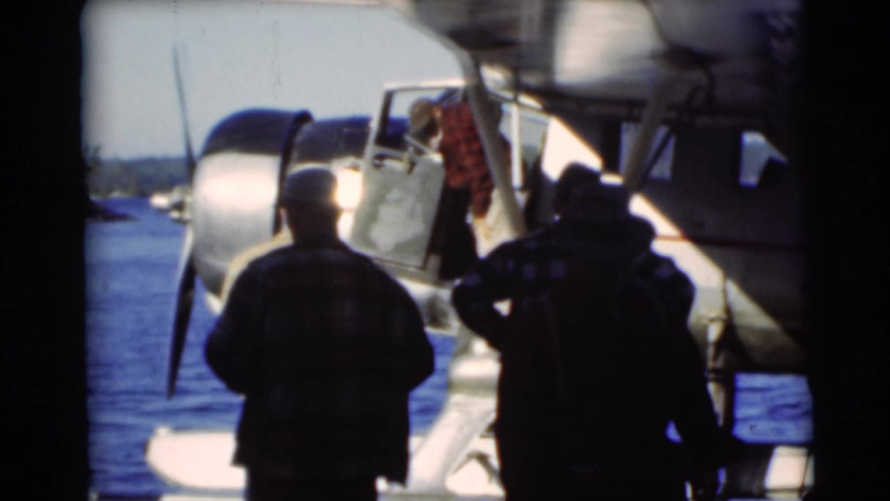 Men stand beside a seaplane at a lakeside dock with blue water behind them.