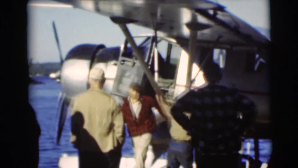 A man in a red plaid shirt and light hat moves near the seaplane door as other men stand by the lakeside in Baraboo footage.