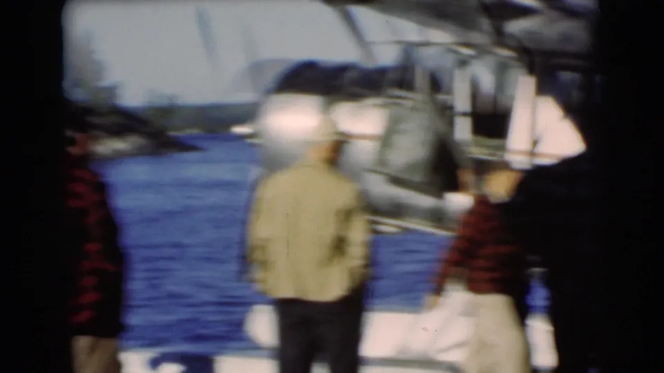 The 8mm frame shows motion blur as men walk near a lakeside seaplane, with blue water and a distant shore behind them.