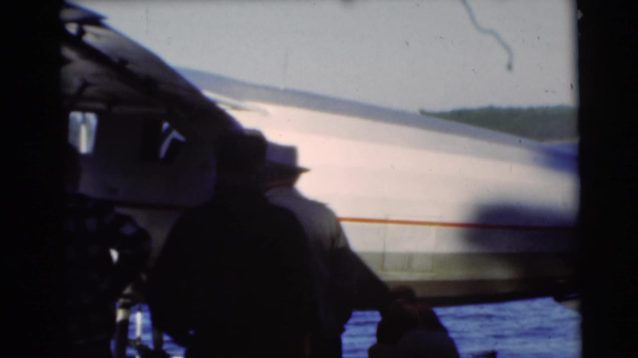 1953 Baraboo-area home-movie frame with silhouetted figures near a pontoon seaplane at the edge of a blue lake.