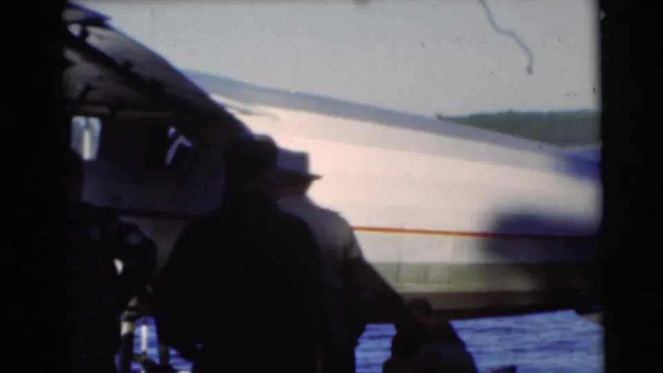 1953 Baraboo-area home-movie frame with silhouetted figures near a pontoon seaplane at the edge of a blue lake.