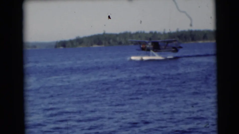 The pontoon seaplane crosses the blue lake in archival 1953 Wisconsin footage, framed by a distant wooded shoreline.