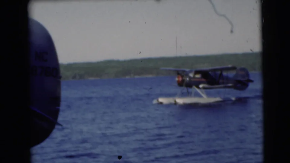A nearby aircraft tail appears at the left edge while the floatplane moves across the lake in Baraboo, Wisconsin footage.