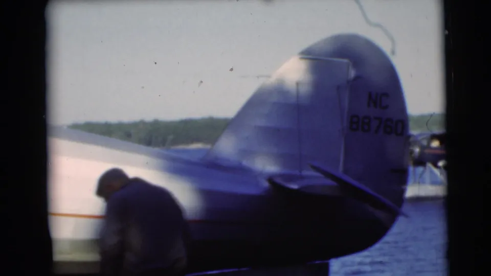 1953 Baraboo, Wisconsin frame with a man near the aircraft fuselage, tail marking visible, and a floatplane on the lake.