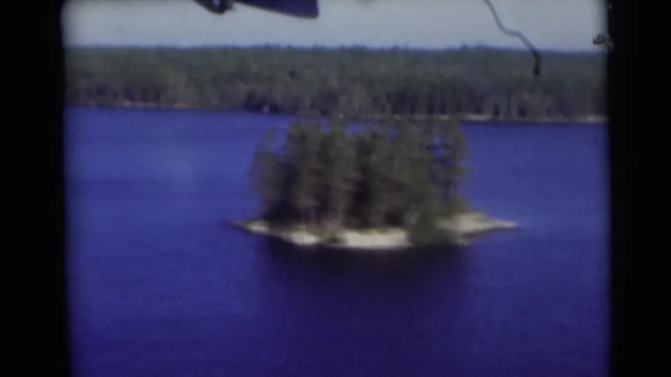 1953 Baraboo, Wisconsin lake view showing a small tree-covered island, blue water, and a wooded shoreline in soft 8mm focus.