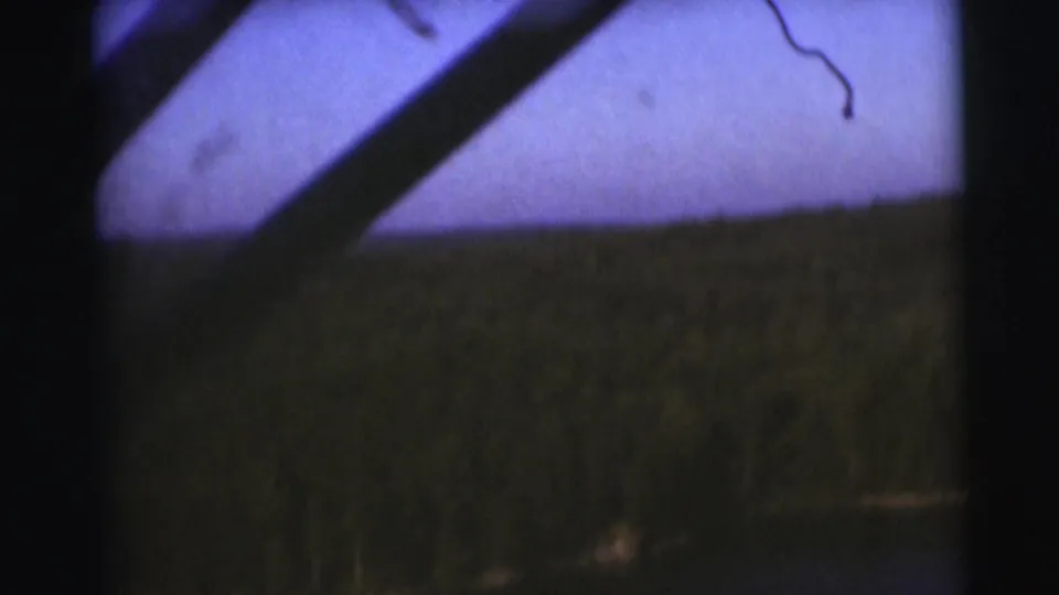 1953 Baraboo, Wisconsin frame with grass near the water, a distant wooded bank, pale sky, and a diagonal dark obstruction at the top.