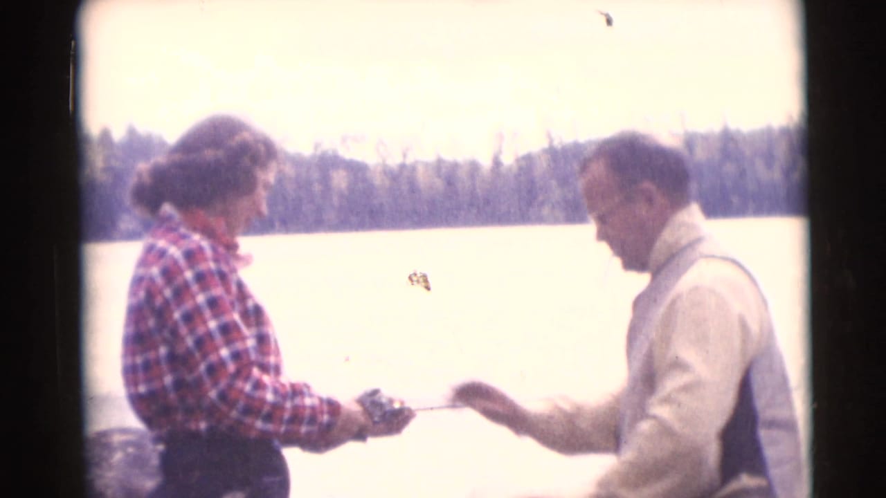 Woman in a plaid shirt and man in a light vest stand beside a lake with fishing gear.