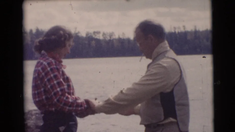 The man appears to guide the woman through a fishing motion at a Baraboo lakeside, with trees visible across the water.