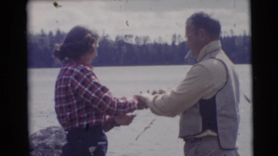 At the lakeside in Baraboo, the woman and man hold fishing gear together, with faded 8mm color and a wooded shoreline behind them.