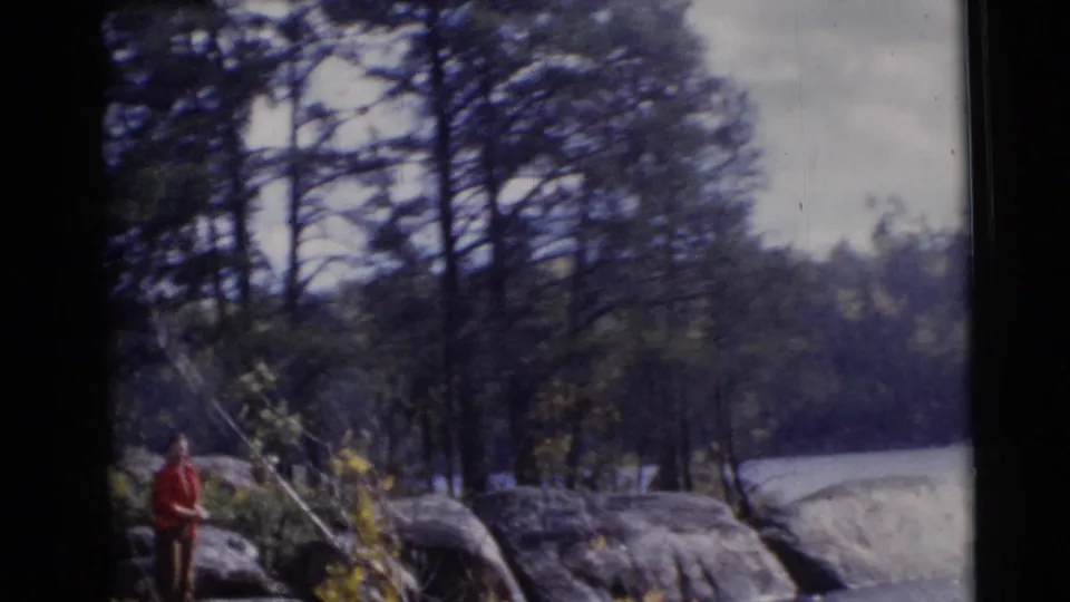 An adult man fishes from a rocky shoreline beside trees and calm water in the Baraboo, Wisconsin home-movie scene.