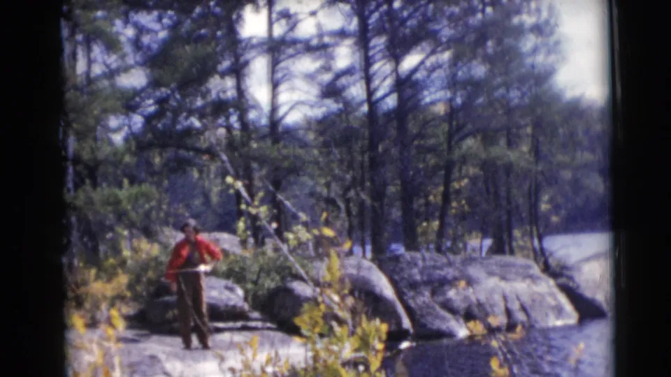The 1953 home-movie frame shows a man fishing on rocks near a wooded Wisconsin lake, with water and shoreline plants in view.