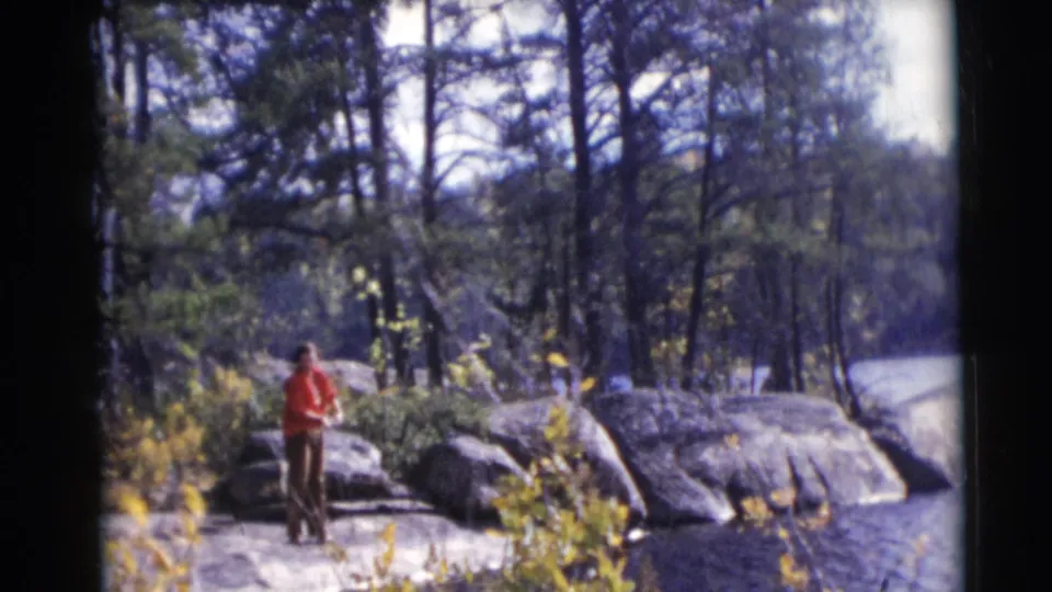 In metadata-supported 1953 Baraboo, Wisconsin footage, a person in a red jacket stands on rocks beside water with trees behind.