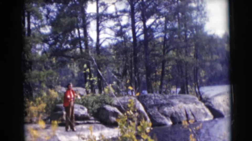 The 1953 Baraboo home-movie frame shows a person in red fishing near large rocks, water, brush, and tall trees.