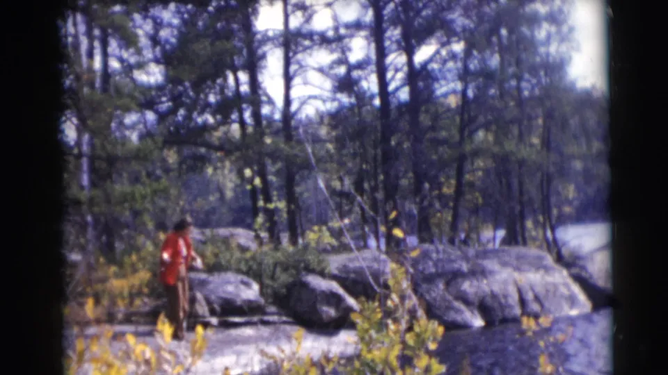 The person leans near the rod on a rocky, tree-lined riverbank in metadata-supported 1953 Baraboo, Wisconsin footage.